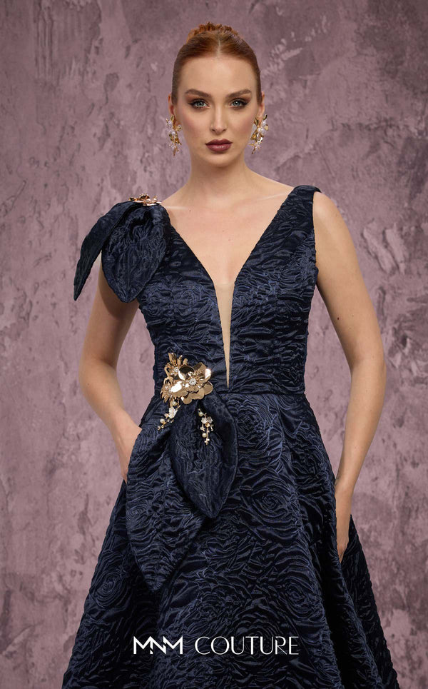 MNM Couture Style Q1150 navy blue brocade ballgown with deep V-neckline, mesh inset, bow detail, and gold and crystal floral embellishments at waist and shoulder. | Couture Shop LA